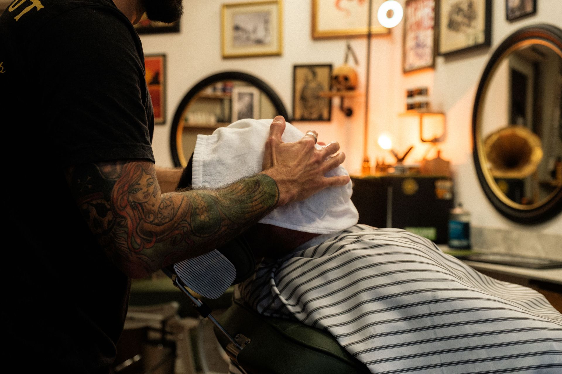 A barber with tattoo sleeves applies a hot towel in a vintage-style barbershop setting.